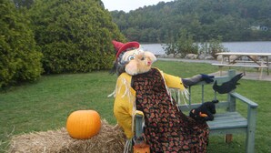A scarecrow with a pumpkin and jack-o'-lantern, a picnic table, and a lake in the background.