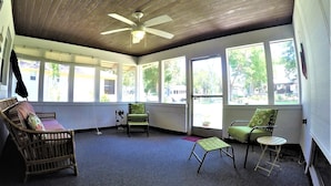 Une véranda avec un ventilateur de plafond, de grandes fenêtres, un canapé en osier, un fauteuil vert et une petite table.