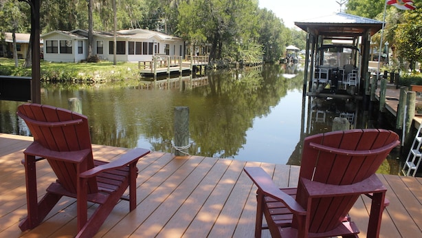 Une terrasse en bois avec deux fauteuils Adirondack donnant sur une rivière paisible, entourée d’arbres et bordée d’un quai surmonté d’une structure couverte.
