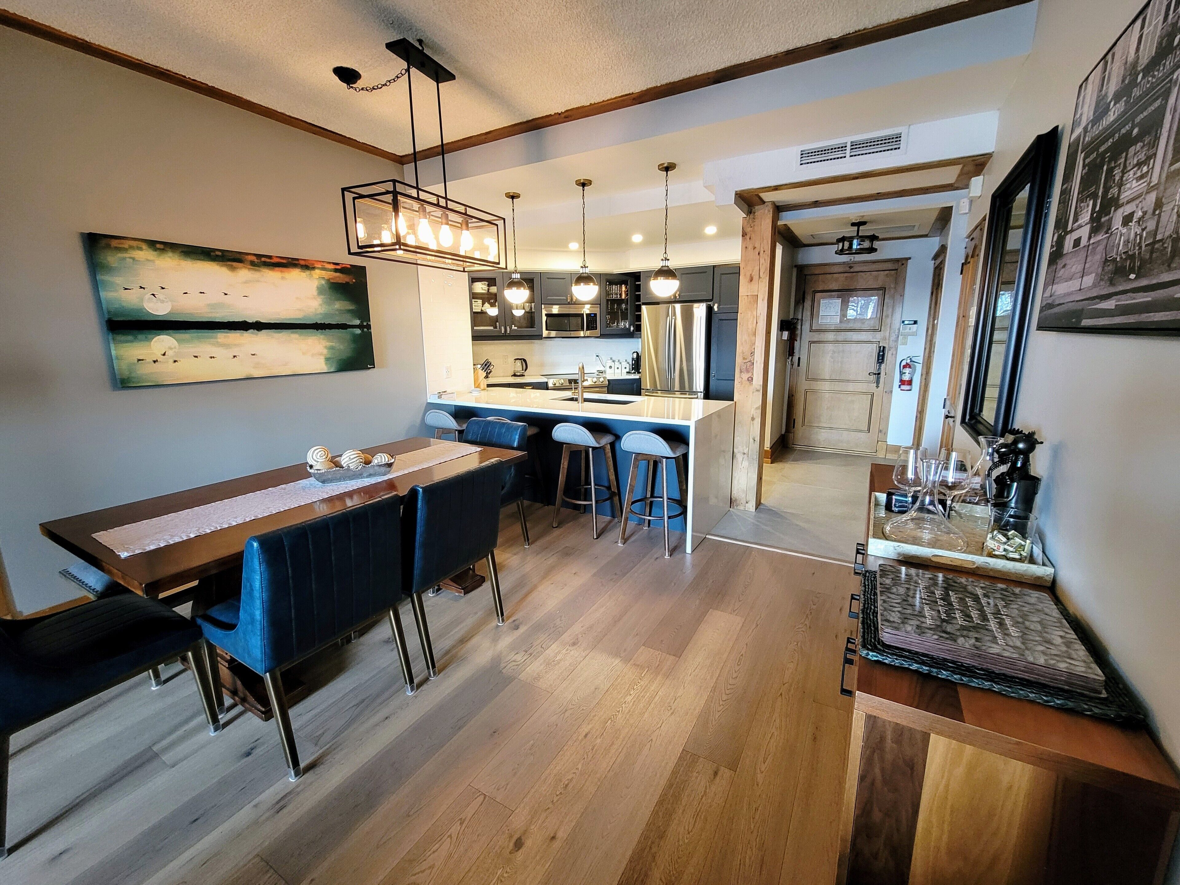 A modern kitchen with a central island, bar stools, pendant lighting, and a dining area with a long table and chairs.