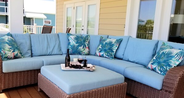 A light blue sectional sofa with tropical leaf-patterned pillows, a wicker ottoman, and a tray with a bottle, glass, and grapes.