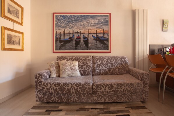 Una sala de estar con un sofá estampado, un cuadro enmarcado de barcos y un comedor con sillas.