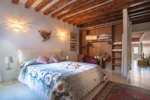 A bedroom with a floral bedspread, wooden ceiling beams, a built-in wardrobe, and a bedside table with a lamp.