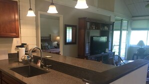 A kitchen with a granite countertop, stainless steel sink, and a built-in oven. There is a dining area with a table and chairs, and a television in the background. - 2 bdrm 2 Bath Condo with Partial Ocean Views, Steps away from Kamaole III Beach  (Kihei)