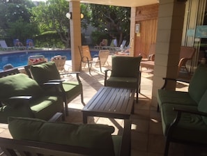 An outdoor seating area with green cushioned chairs and a wooden table, overlooking a pool and greenery.
