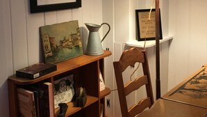 A dining area with a wooden table, a chair, a lamp, a shelf with books and a painting, and a framed picture on the wall.