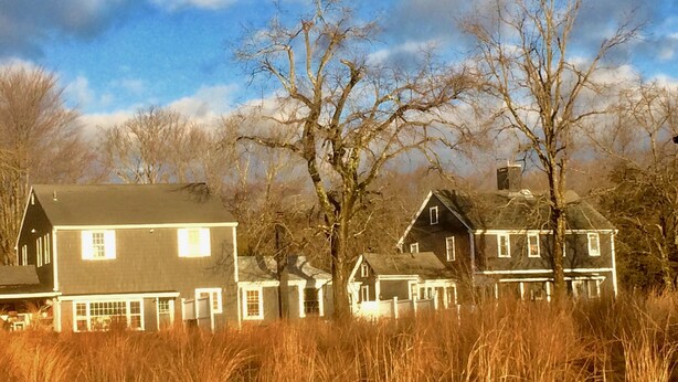 A large, two-story house with a dark roof and white trim, surrounded by tall grass and trees.