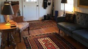 A living room with a sofa, a chair, a table with a lamp, a coat rack with hats, and a door.