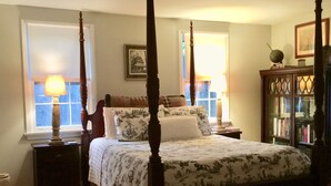 A four-poster bed with a floral bedspread, two bedside tables with lamps, a nightstand, a bookshelf, and a window with a view.