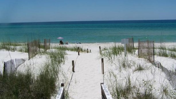 Un sentiero sulla spiaggia che conduce verso l'oceano, delimitato da una recinzione su entrambi i lati, con una persona seduta sotto un ombrellone.