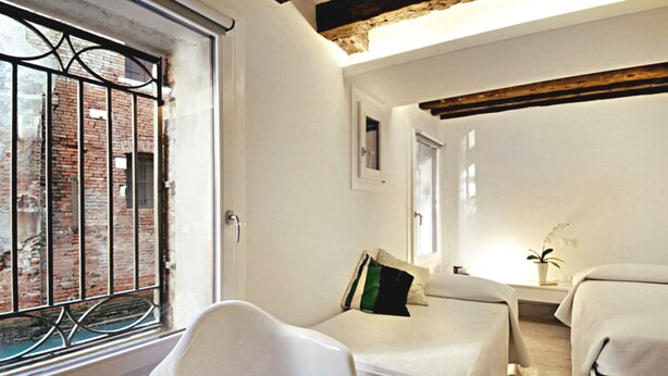 A hotel room with two beds, a window with a metal grill, a white chair, and a small table with a plant. - Charming, views Of Canals, Gondolas, Church Of The Miracoli (Venezia)