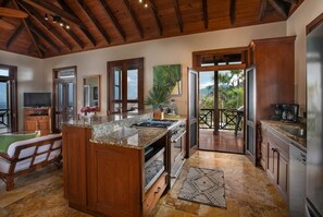 A spacious kitchen with wooden cabinets, granite countertops, and a large island. There is a dining area with a table and chairs, and a view of palm trees through the open door.