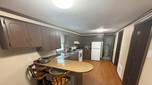 A kitchen with wooden cabinets, a granite countertop, and a built-in stove. There are two bar stools and a refrigerator. - Wendell’s Place on Red Creek - Uncle Ralph’s Cabin Rentals - PRIVATE BEACH (Perkinston)