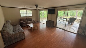 A spacious living room with a large sofa, a coffee table, and a television. There is a sliding glass door leading to an outdoor deck with patio furniture. - Wendell’s Place on Red Creek - Uncle Ralph’s Cabin Rentals - PRIVATE BEACH (Perkinston)