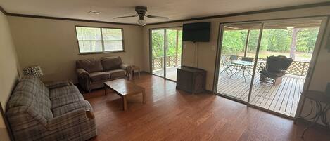 A spacious living room with a large sofa, a coffee table, and a television. There is a sliding glass door leading to an outdoor deck with patio furniture.