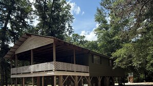 A rustic wooden pavilion with a covered porch and lattice railing, surrounded by trees and a gravel path. - Wendell’s Place on Red Creek - Uncle Ralph’s Cabin Rentals - PRIVATE BEACH (Perkinston)