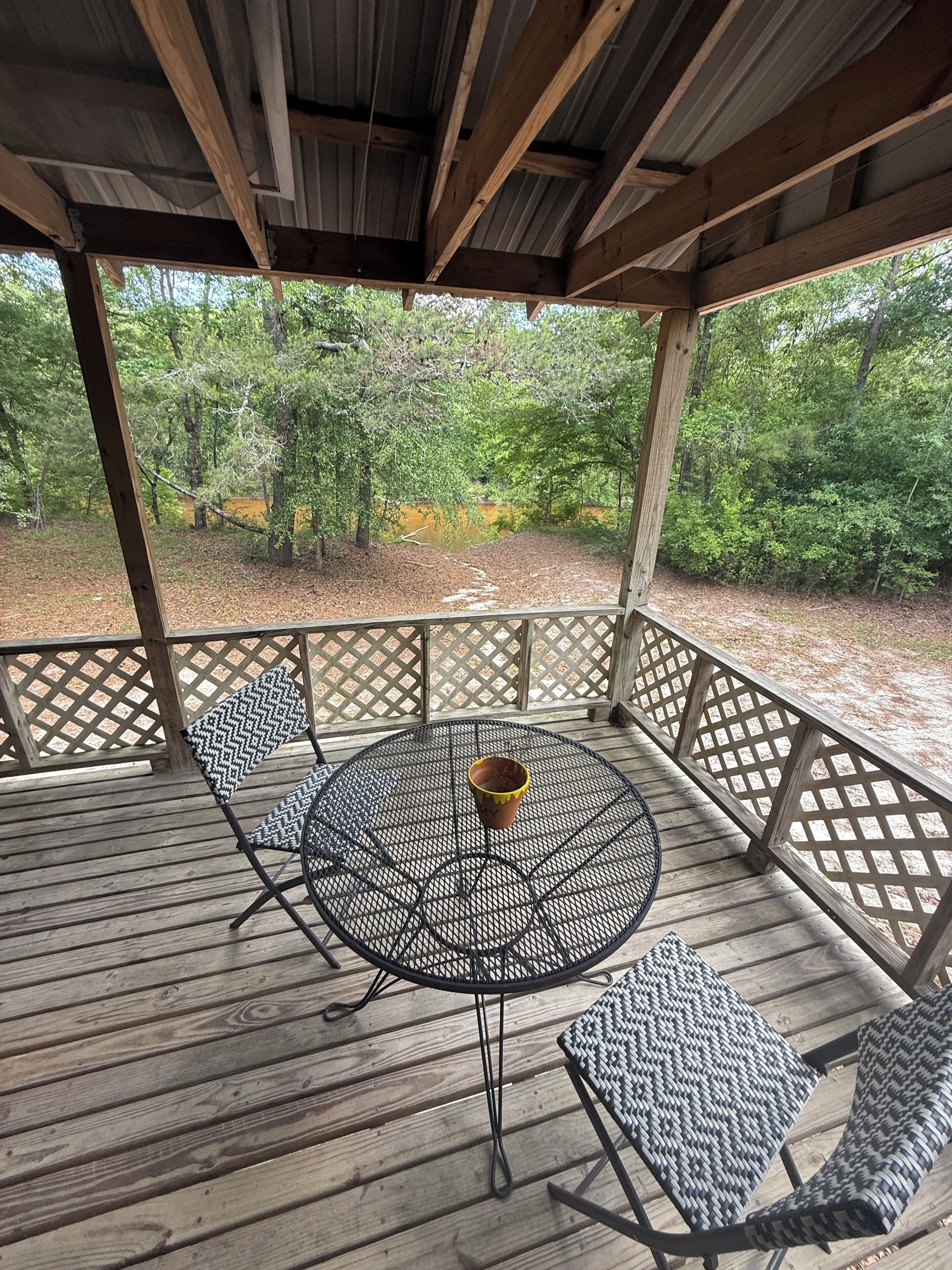 A covered outdoor seating area with a round metal table and two patterned chairs, surrounded by a wooden deck and lattice railings, overlooking a wooded area.