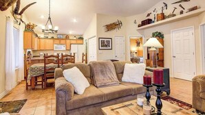 A cozy living room with a sofa, a coffee table, and a fireplace. There is a dining area with wooden furniture and a kitchen in the background.