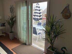 A room with a sliding glass door leading to an outdoor patio with a table and chairs. There are potted plants on either side of the door, a ship's wheel decoration on the wall, and a small shelf with books and a lamp. - Just 1 block from the boardwalk, beach, and water park! Book now for 2026! (Wildwood)