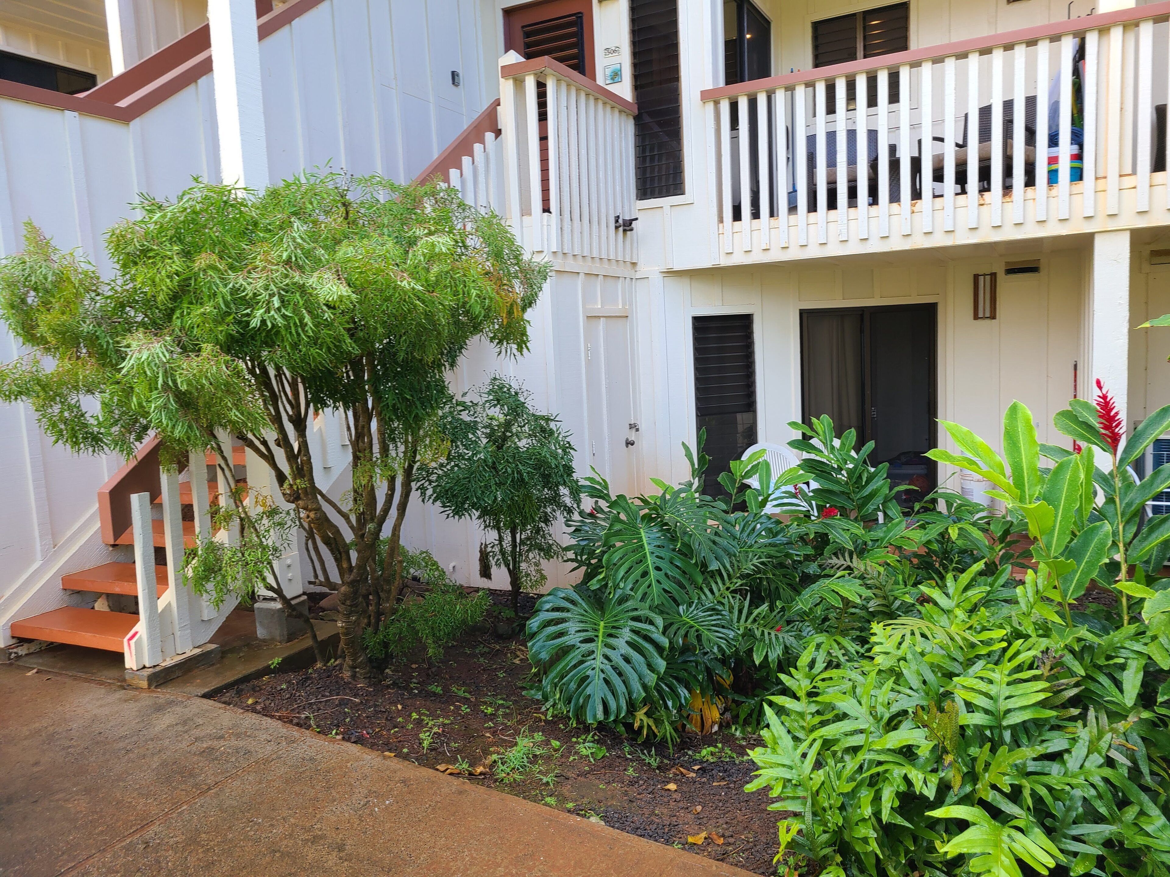 A residential building with a staircase leading to a balcony, surrounded by various plants and a small garden area.
