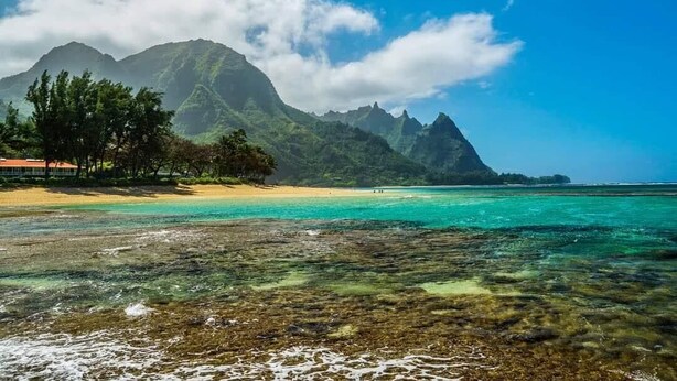 Ein Strand mit klarem türkisblauem Wasser, sandigem Ufer und einem Berg im Hintergrund.