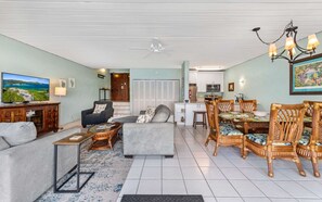 A living room with a sofa, armchair, coffee table, dining table with chairs, a TV, and a kitchen area.