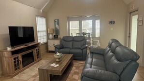 Un salon spacieux avec un haut plafond, un grand téléviseur à écran plat, un canapé d’angle et une table basse. Il y a deux fenêtres équipées de stores, une porte avec un panneau indiquant la sortie, et une petite table ornée d’objets décoratifs.