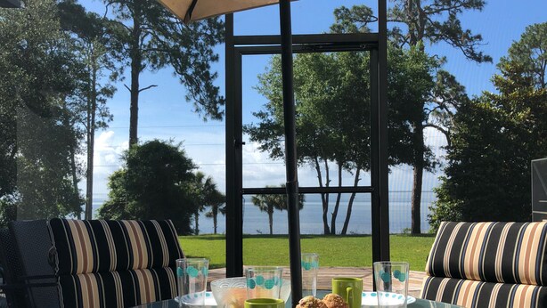Une table en verre avec des coussins à rayures, un parasol beige et une vue sur les arbres et l’eau.