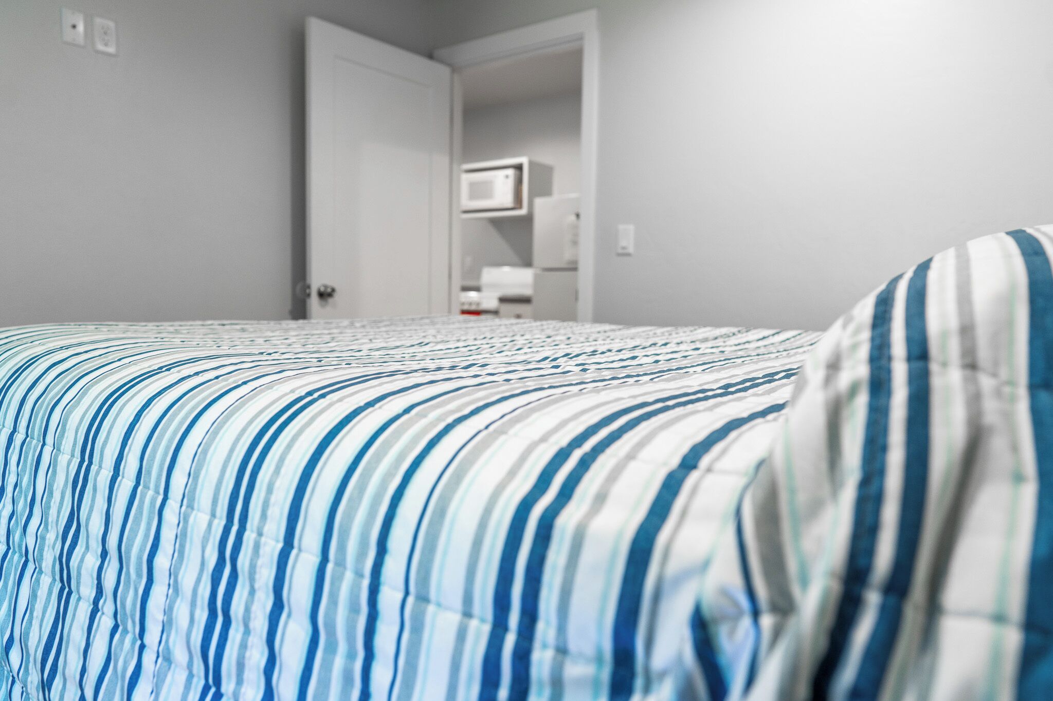 A bedroom with a striped bedspread, a door leading to a kitchen area, and a microwave visible in the background.