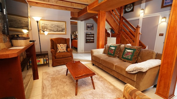Een knus woongedeelte met een houten trap, een bruine bank met decoratieve kussens, een houten salontafel en een lijst met een landschapschilderij aan de muur.