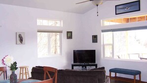 A living room with a ceiling fan, a TV, a couch, a bench, and a dining table with chairs. There are windows with blinds and a picture frame on the wall. - You have to get out-of-town to see the wildlife (Homer)