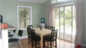 A dining area with a wooden table and six chairs, a window with a view of greenery, and a sliding glass door leading outside. - Cedar Cottage - Kelleys Island (Lake Erie) Vacation Retreat (Kelleys Island)