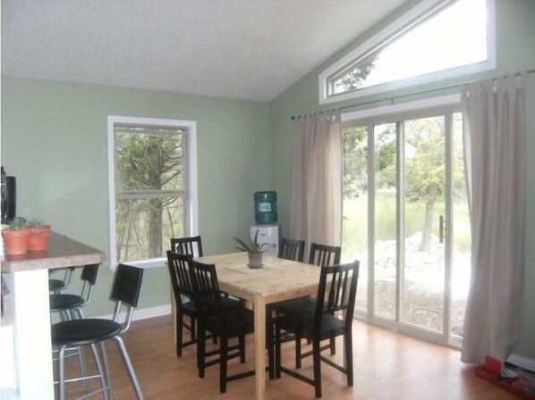 A dining area with a wooden table and six chairs, a window with a view of greenery, and a sliding glass door leading outside.
