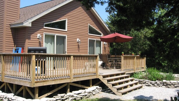 A wooden deck with a stone wall, outdoor furniture, and a red umbrella. - Cedar Cottage - Kelleys Island (Lake Erie) Vacation Retreat (Kelleys Island)