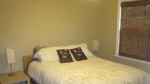 A neatly made bed with white bedding and two decorative pillows, a bedside table with a lamp, and a window with blinds. - Cedar Cottage - Kelleys Island (Lake Erie) Vacation Retreat (Kelleys Island)