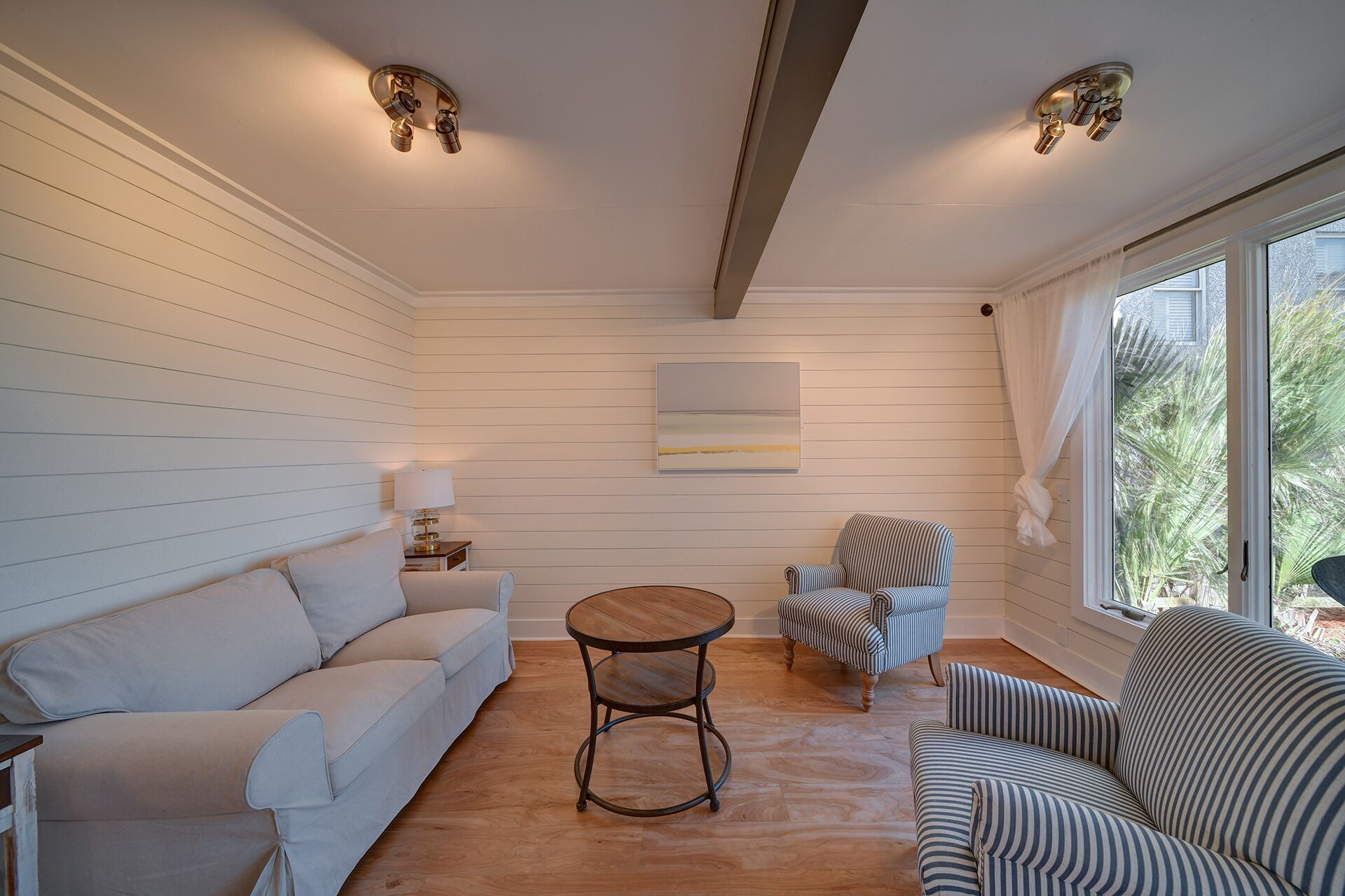 A room with a white paneled wall, a wooden floor, a white sofa, a striped armchair, a round wooden table, and a window with a view of greenery.