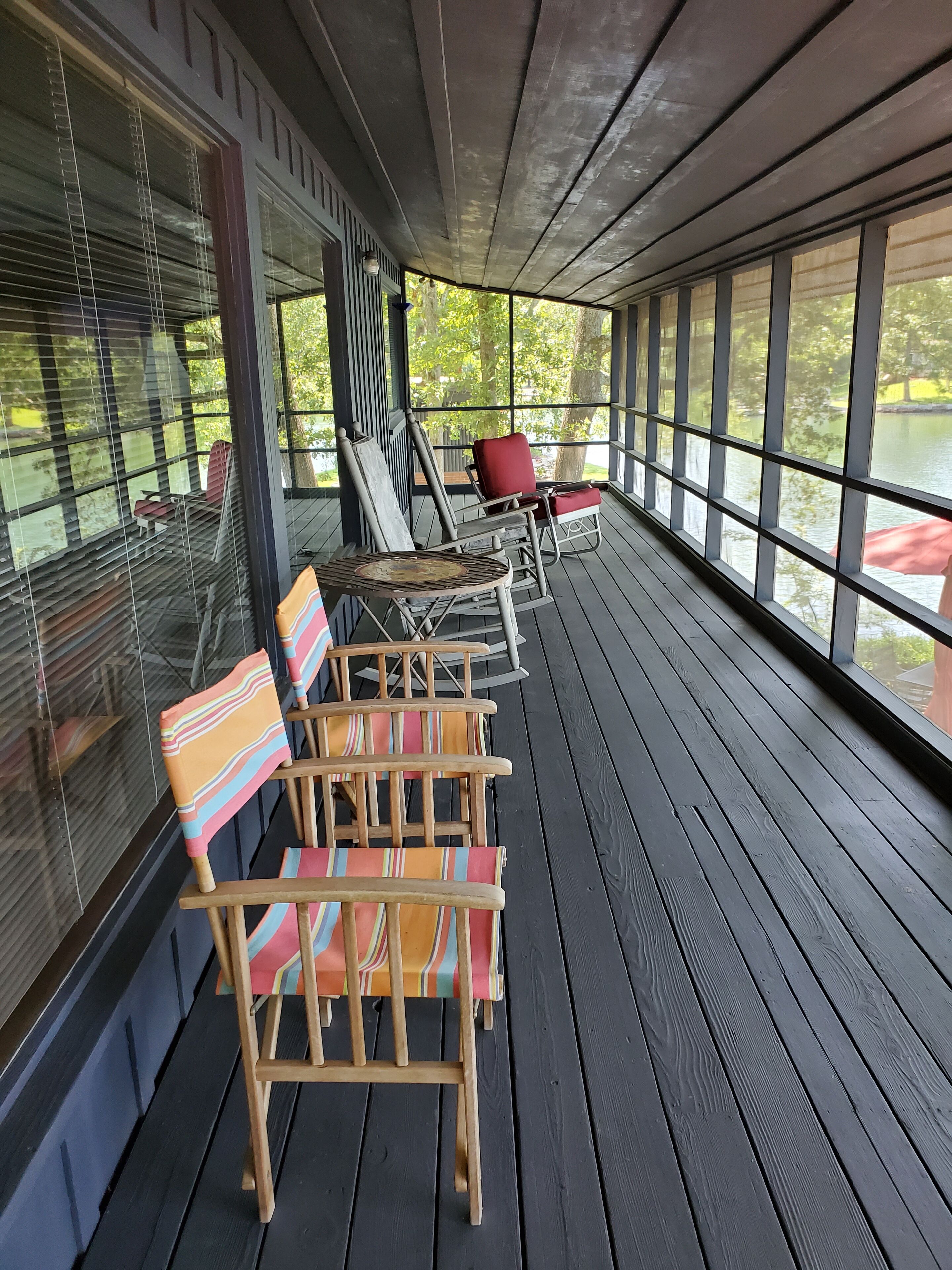 Un área de estar al aire libre techada, con sillas y mesa de madera, ventanas amplias y vista a un cuerpo de agua.