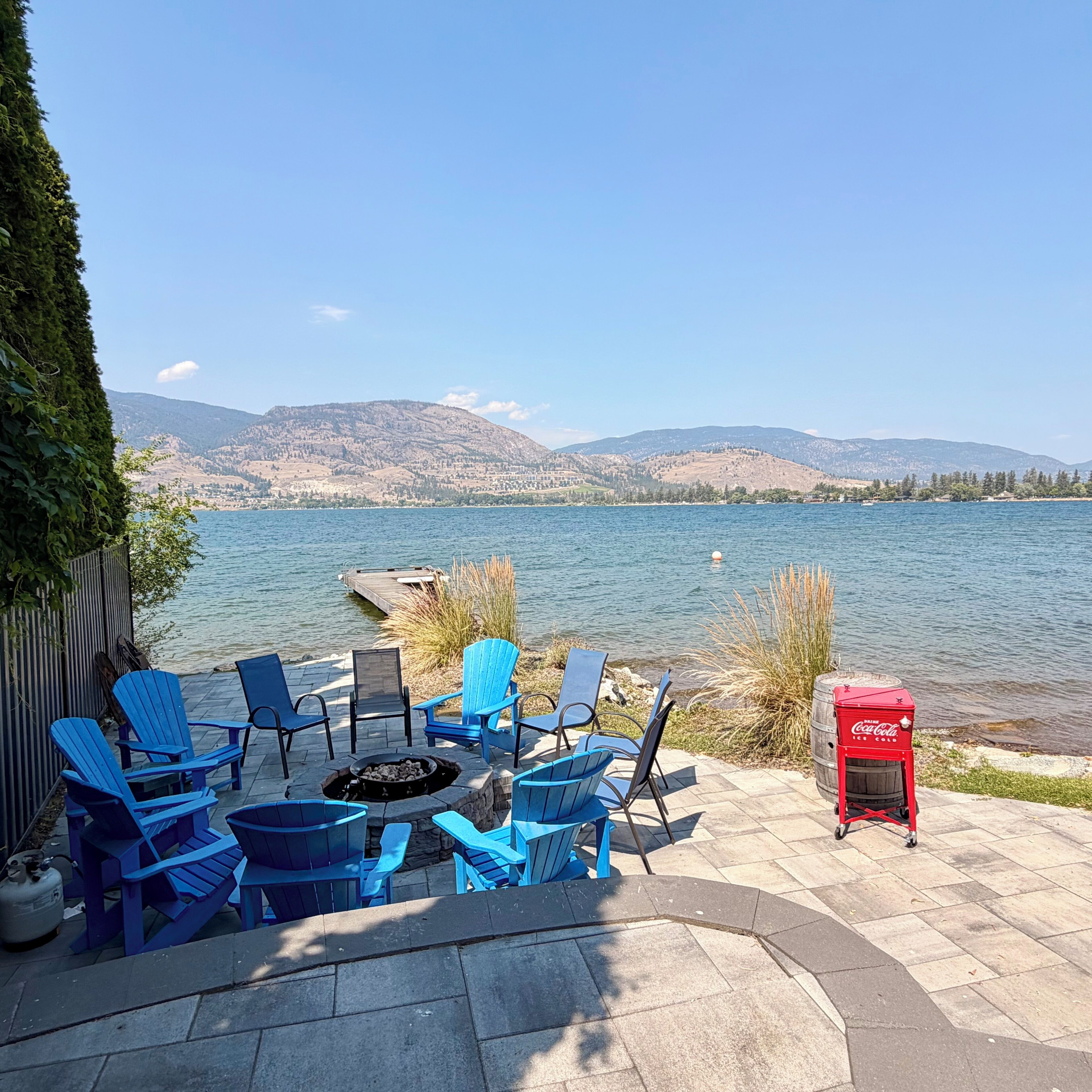 A lakeside patio with blue Adirondack chairs, a fire pit, and a red Coca-Cola cooler.