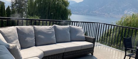 Een balkon met een rieten bank en kussens, met uitzicht op een meer en bergen.