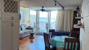 A spacious living area with a wooden floor, a dining table with chairs, a sofa, and a view of the sea through large windows. - Spacious one bedroom condo on a white sandy beach! (Hauula)