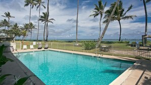 A clear blue pool with lounge chairs and palm trees, surrounded by a fence, with a view of the ocean. - Spacious one bedroom condo on a white sandy beach! (Hauula)