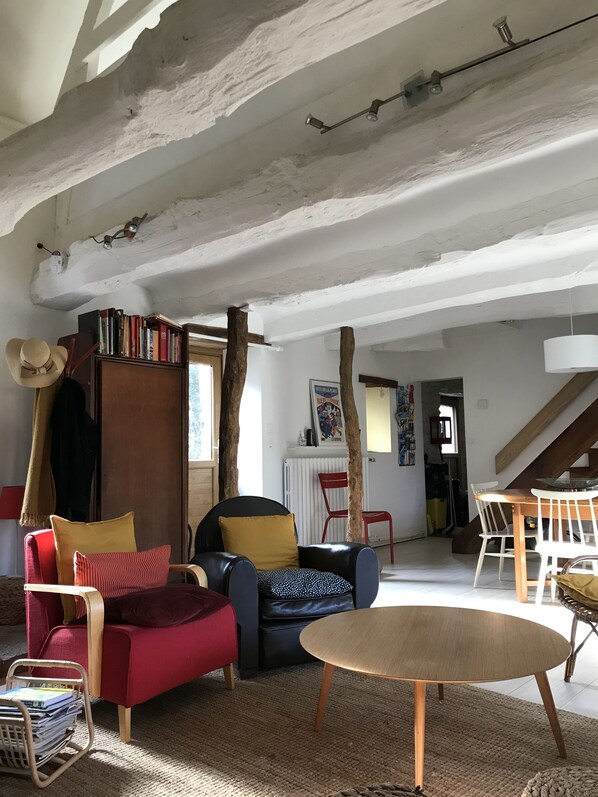 Uma sala de estar acolhedora com pé-direito alto, vigas de madeira rústicas, uma estante, uma poltrona vermelha, uma poltrona preta e uma mesa de madeira redonda.