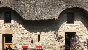 Een gebouw van steen met rieten dak, twee rode stoelen en een tafeltje ervoor.