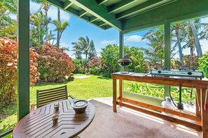 Un espace extérieur abrité, équipé d’une table et de chaises, d’un barbecue et offrant une vue sur un jardin tropical.