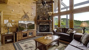 Un salon chaleureux avec une cheminée en pierre, des murs en bois et de grandes fenêtres donnant sur l’extérieur.