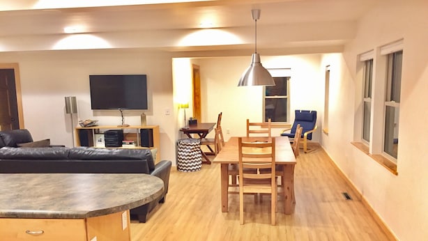 Una sala de estar moderna con un sofá, una mesa de comedor con sillas, un televisor montado en la pared y una lámpara de techo.