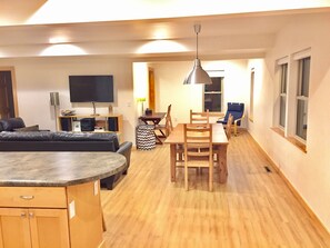 A modern living room with a sofa, a dining table with chairs, a TV mounted on the wall, and a ceiling light.
