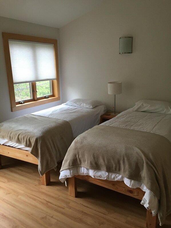 Two single beds with wooden frames and white bedding, a window with blinds, a bedside lamp, and a small wall-mounted light fixture.