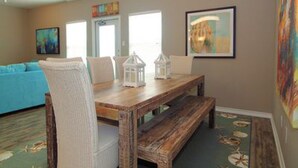 Un comedor con una mesa de madera, sillas blancas y un banco. Hay dos ventanas con cortinas, un cuadro en la pared y una alfombra colorida en el suelo.