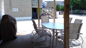 Een overdekt terras buiten met een tafel en stoelen, een barbecue en een trap naar een terras.